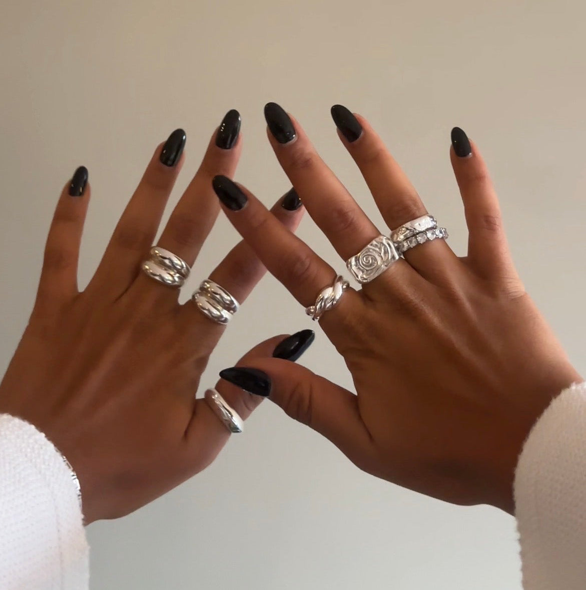 Two hands with black nail polish and silver rings on a neutral background