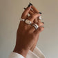 A close-up of a person's hand wearing multiple silver rings, including a prominent rectangular signet-style ring and stacked bands, against a plain beige background.