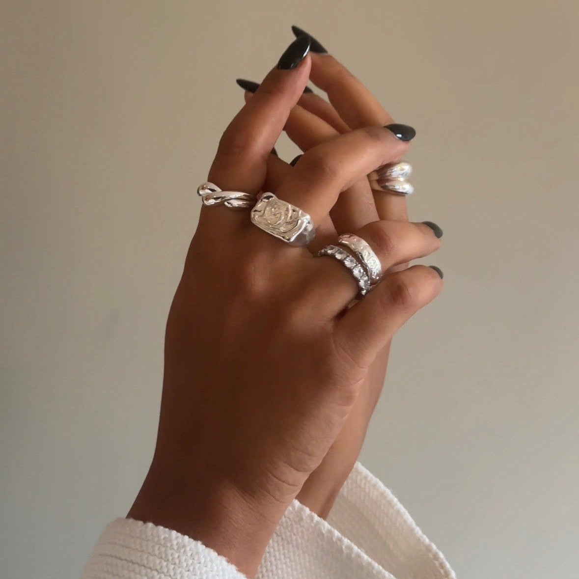 A close-up of a person's hand wearing multiple silver rings, including a prominent rectangular signet-style ring and stacked bands, against a plain beige background.